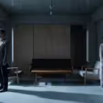 A couple standing far apart in a dimly lit, modern living room, looking away from each other.