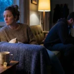 A couple sits on a sofa in a dimly lit room, one person looking at a phone while the other looks away, capturing a moment of secrecy.