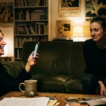 A woman laughs at her phone while her friend sits beside her on a sofa with a forced, thin smile and envious eyes.