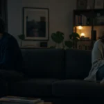 A couple sits on opposite ends of a long sofa in a dimly lit living room, staring away from each other with a large physical gap between the