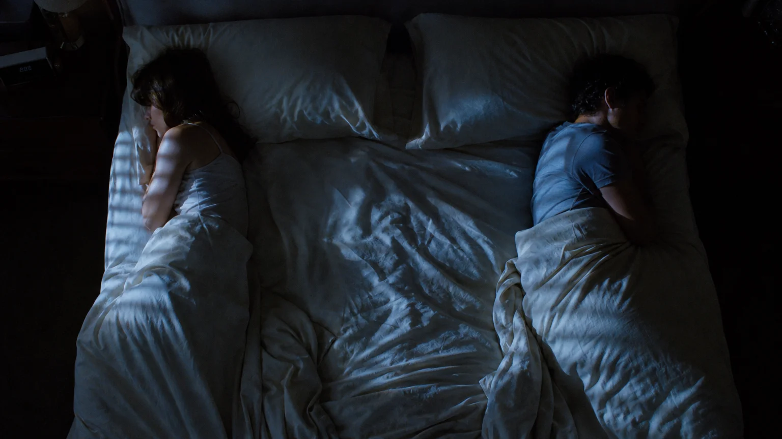 Two people lie on the absolute edges of a large bed, facing away from each other, separated by a wide expanse of empty white sheets.