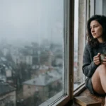 An adult in a knit sweater sits on a window sill on a gray day, looking out over a misty city with a steaming mug.