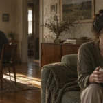 A woman holding a mug sits pensively on a couch while a man sits at a dining table facing away in the background.
