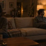 A man sits on a sofa looking out a rainy window while a woman sits facing away in a dim living room.