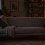 A man and woman sit at opposite ends of a long sofa in a dimly lit living room, looking away from each other.