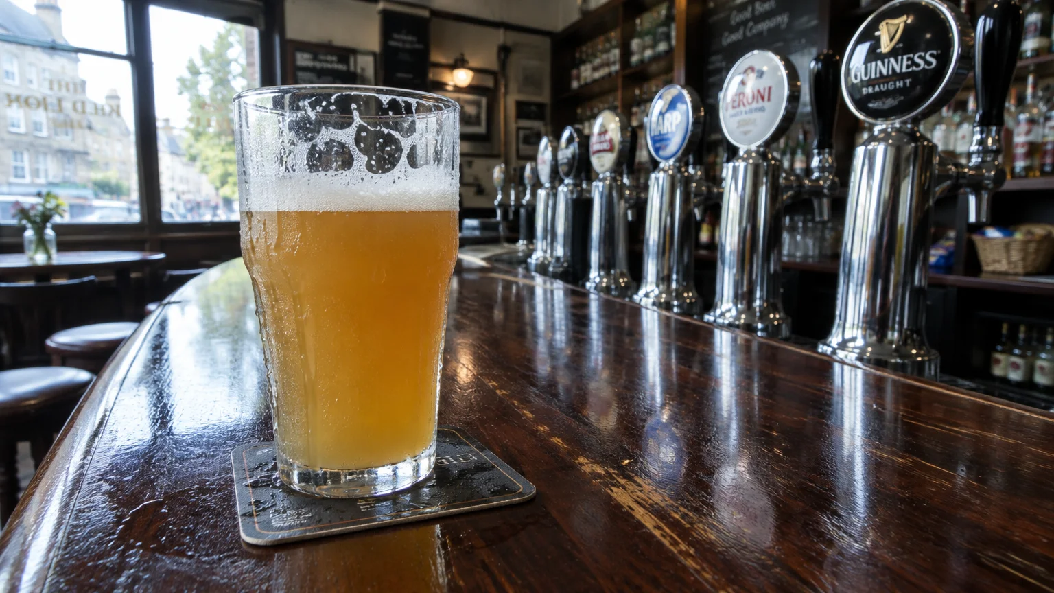 A pint of light beer sits on a wooden pub counter in front of a row of chrome draft beer taps.