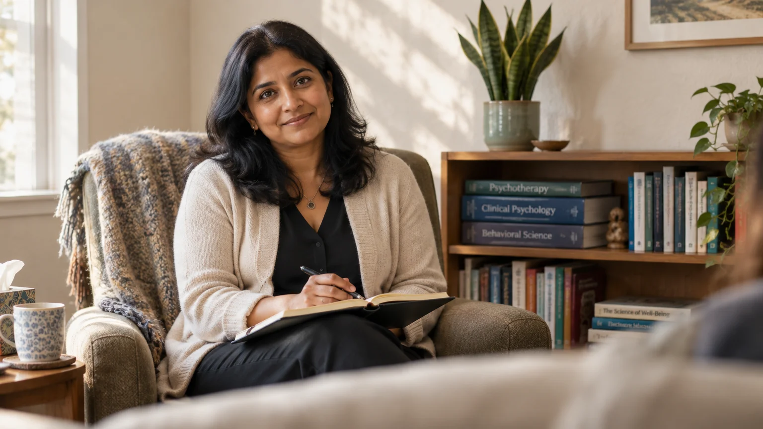 A smiling therapist sits in an armchair holding a notebook and pen in an office with psychology books on a shelf.