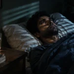 A man lies awake in bed in a dark room, staring upward beside a digital clock showing 3:00.