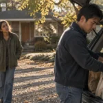 A man unloads groceries from an open car trunk while a woman watches him in a gravel driveway outside a house.