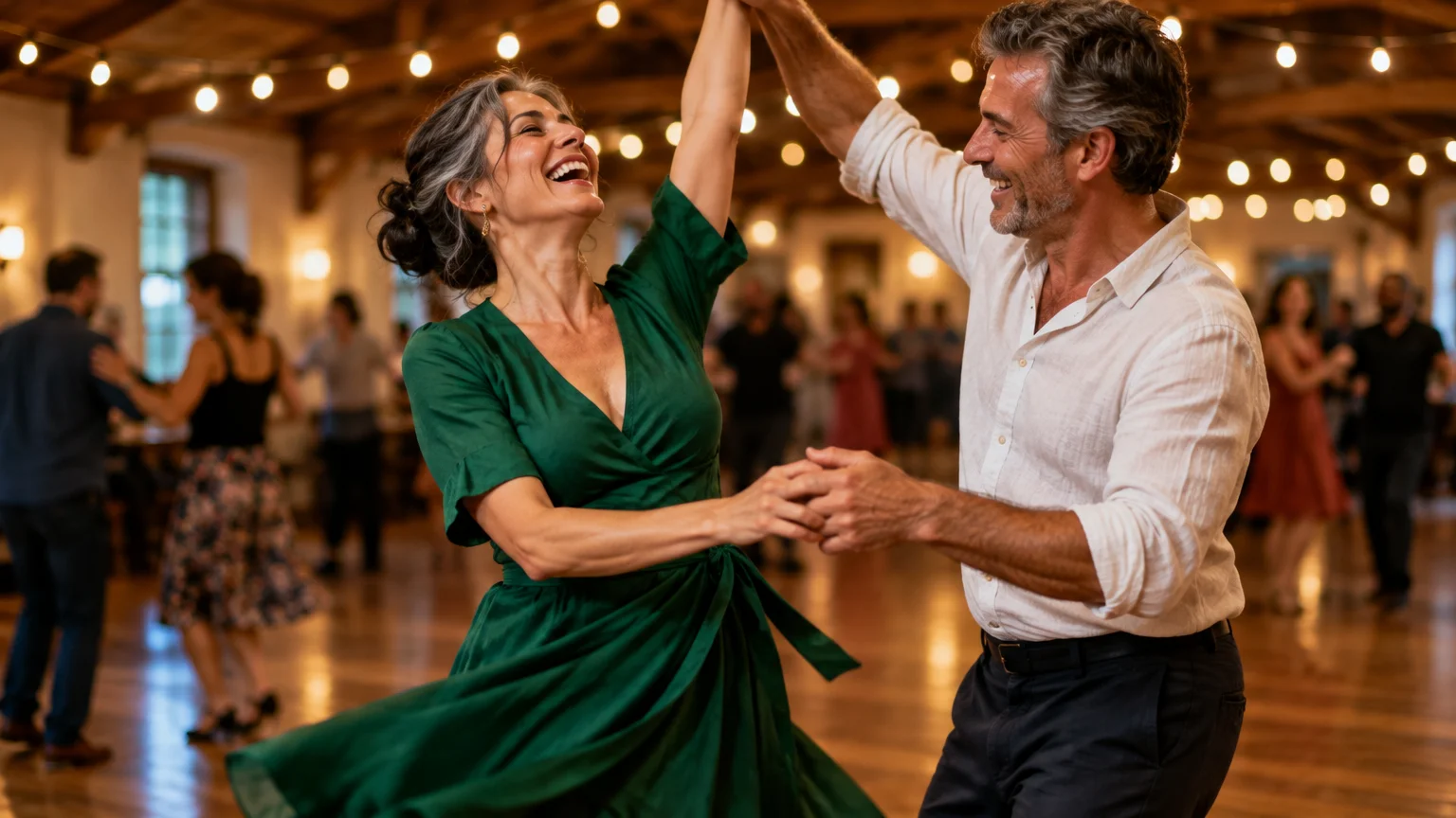 A smiling mature couple dances together on a wooden floor in a warmly lit hall decorated with string lights.