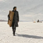 A man carrying wooden shutters walks across a cracked desert plain away from a figure seated near a campfire.
