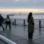 Woman in a trench coat standing on a wet waterfront ferry pier overlooking the water as commuters walk past.